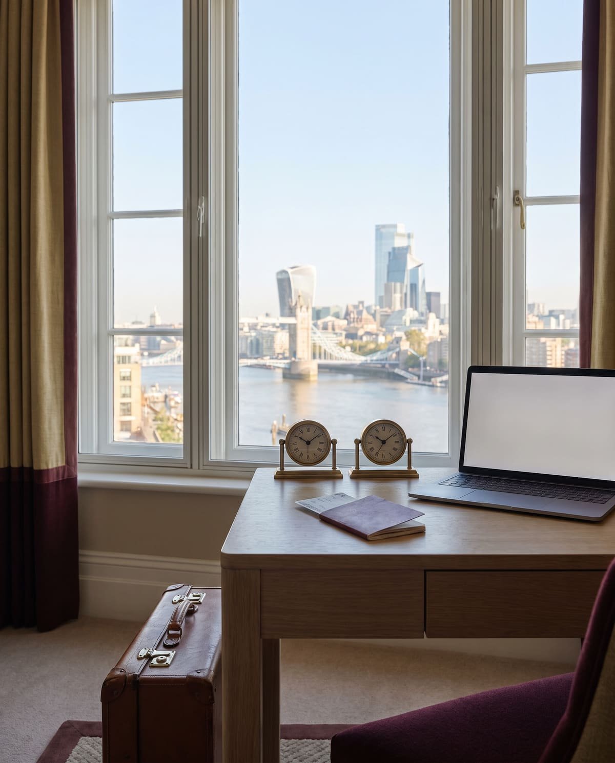 A desk by a window overlooking Tower Bridge and the City of London, with a passport, a laptop, two clocks set to different times and a suitcase